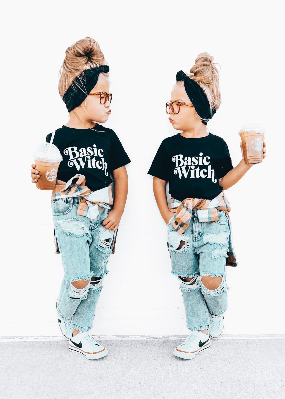 Two children wearing matching outfits with 'Basic Witch' shirts and ripped jeans, holding drinks.
