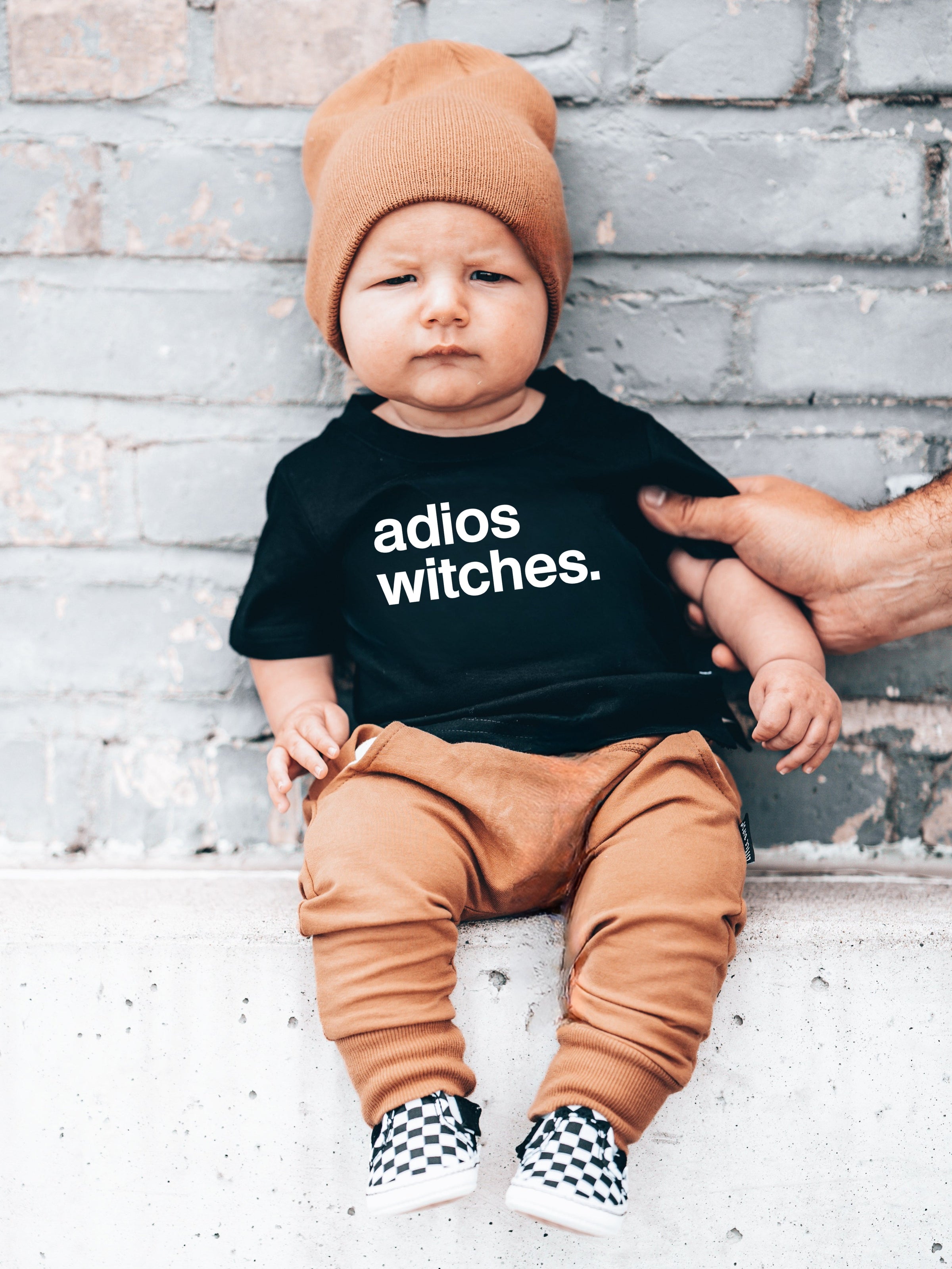 Baby wearing a black t-shirt with 'adios witches.' text, sitting on a brick wall.