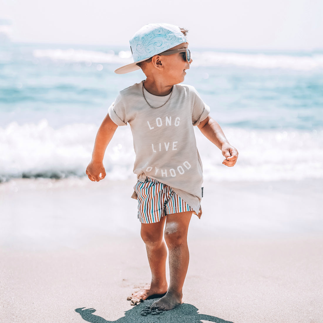 Child standing on a sandy beach wearing a cap and sunglasses with ocean in the background