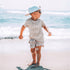 Child standing on a sandy beach wearing a cap and sunglasses with ocean in the background