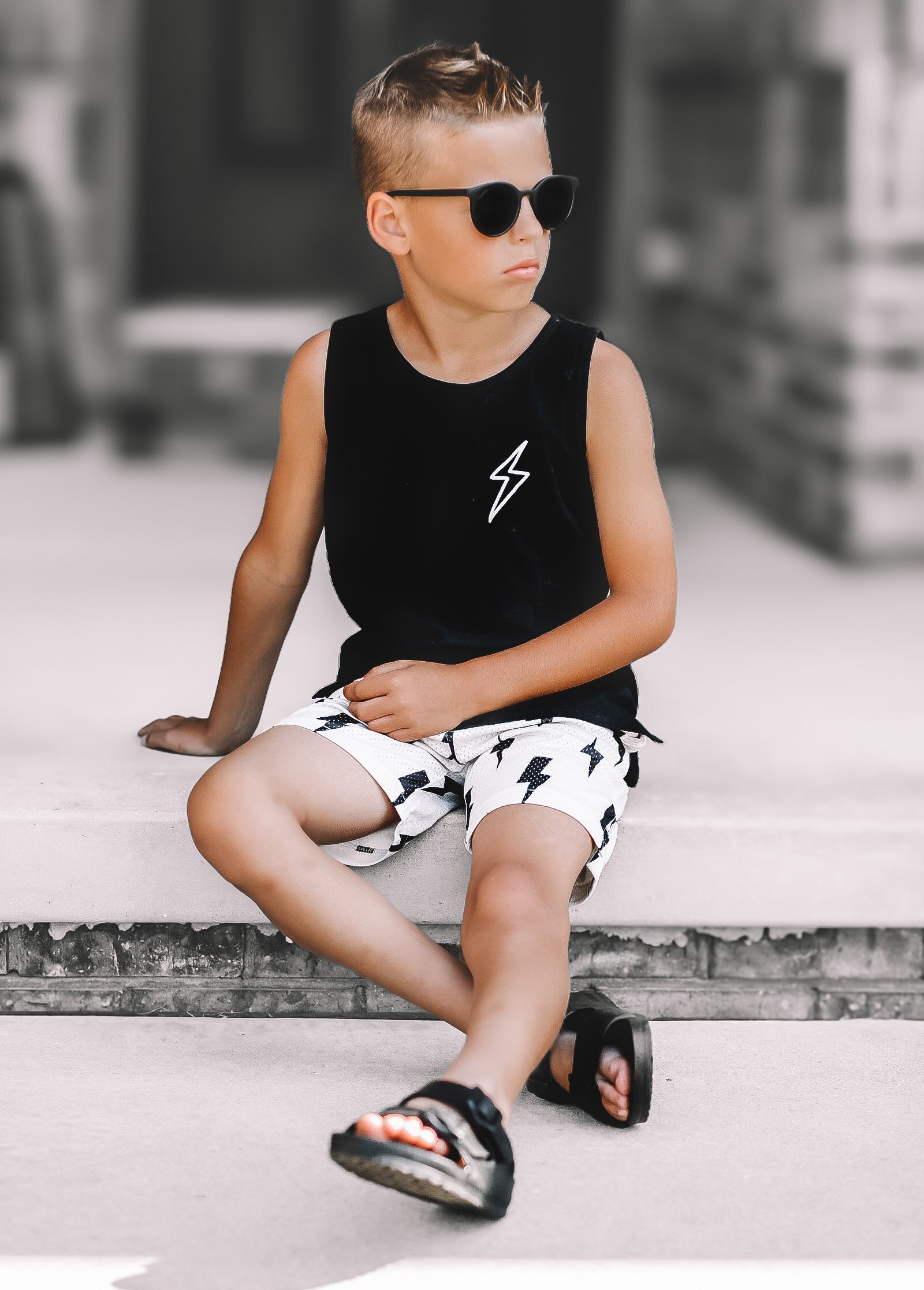 Child wearing a black tank with bolt graphic and white shorts with black bolts, sitting on a concrete surface.