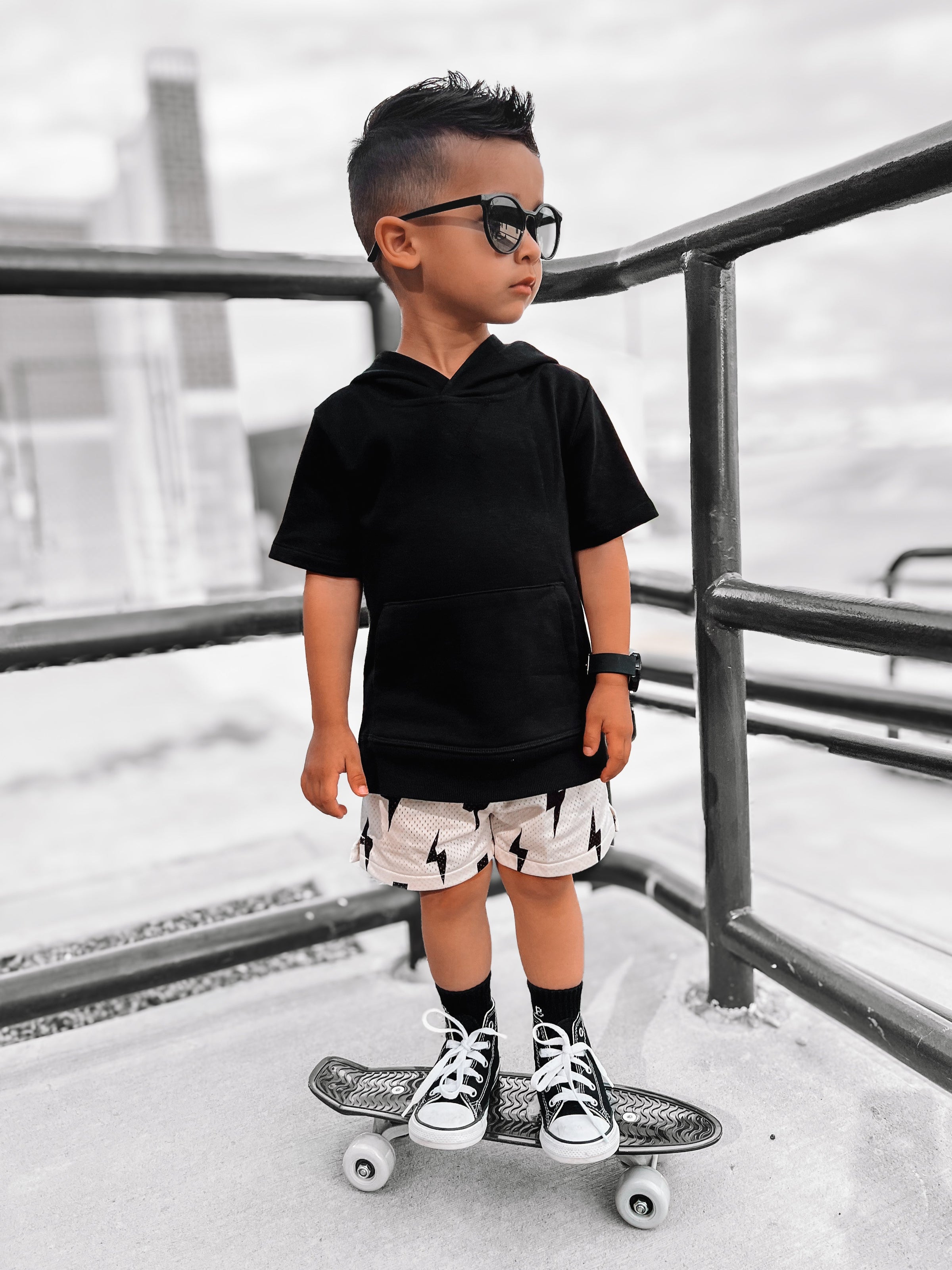 Child wearing sunglasses and a black outfit with a skateboard on a concrete surface