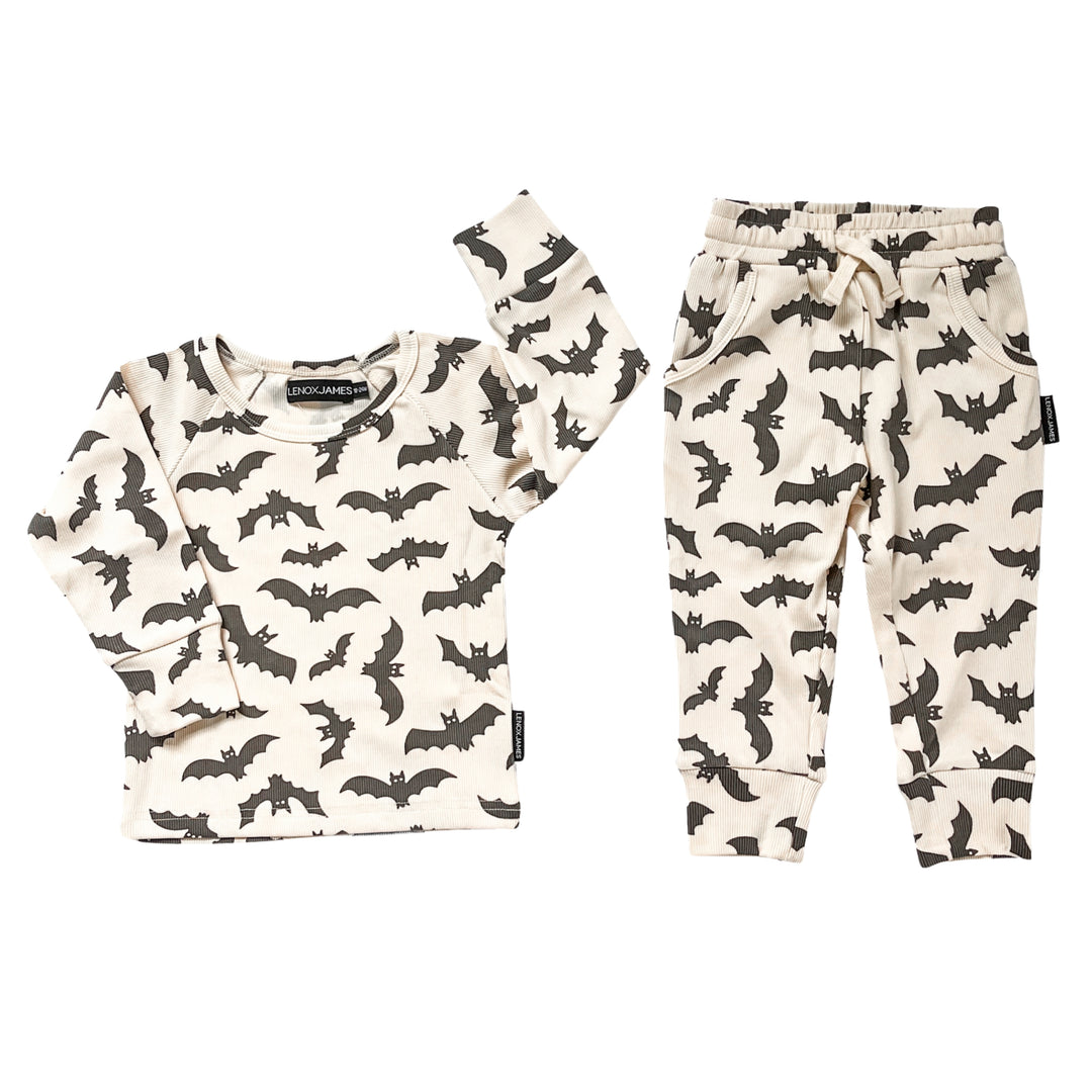 A two-piece children's outfit set featuring a long-sleeve top and matching jogger pants, both in an off-white color with an all-over black bat print. The top has a crew neck and long sleeves, while the pants have an elastic waistband with a drawstring and side pockets.