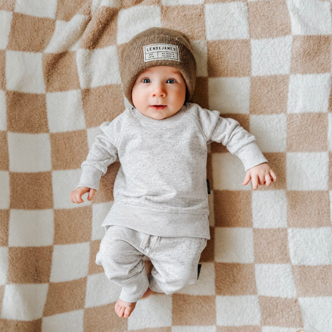 Baby wearing LENOX JAMES ash gray joggers with a matching crewneck sweatshirt and brown knit beanie, lying on a beige and cream checkered blanket. Coordinating baby sweatsuit outfit.