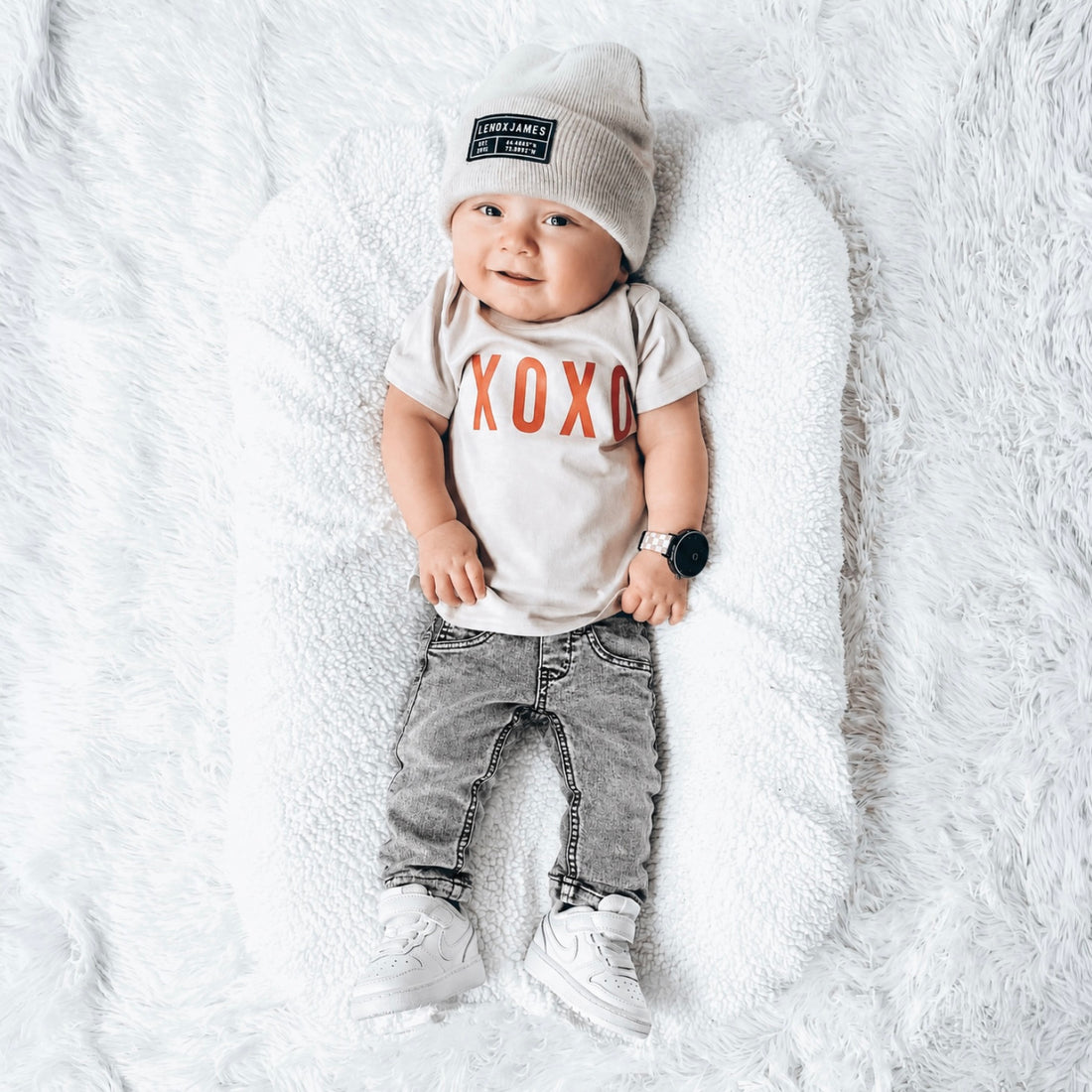 Baby boy wearing LENOX JAMES classic gray wash denim jeans, an XOXO graphic tee, and a knit beanie while lying on a soft white blanket. Features classic non-distressed gray denim with a comfortable everyday fit.