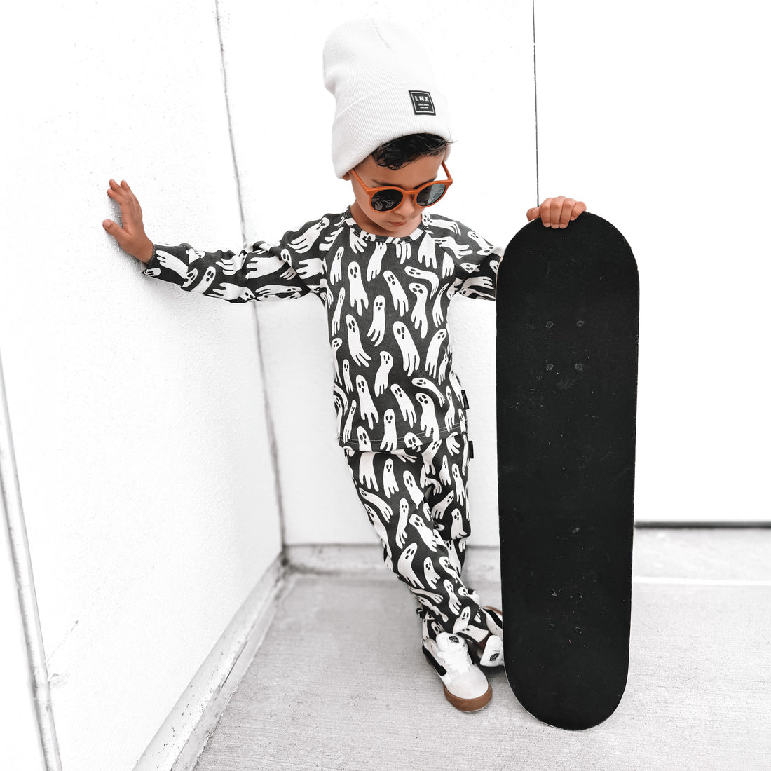 A child wearing a black and white ghost-patterned long-sleeve top and matching pants, a white beanie, and orange-tinted sunglasses, leaning against a white wall while holding a black skateboard.