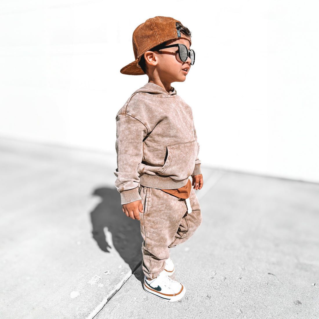 Toddler boy wearing LENOX JAMES tan acid wash joggers with a matching vintage-wash hoodie, brown snapback hat, sunglasses and white sneakers for a surf and skate-inspired kids&