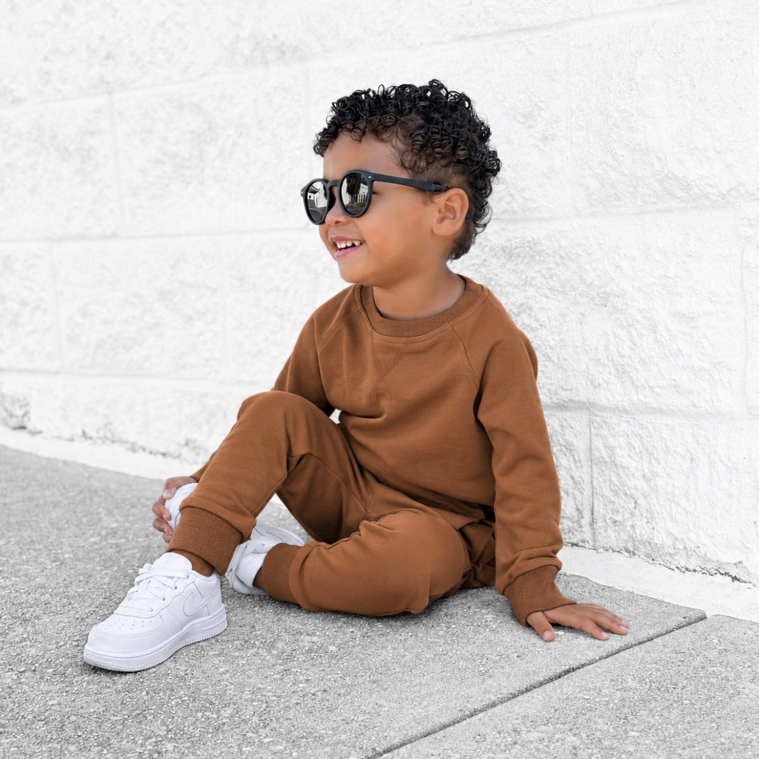 LENOX JAMES terracotta joggers styled on a toddler boy with a matching brown crewneck sweatshirt, black sunglasses and white sneakers for a cool everyday outfit.