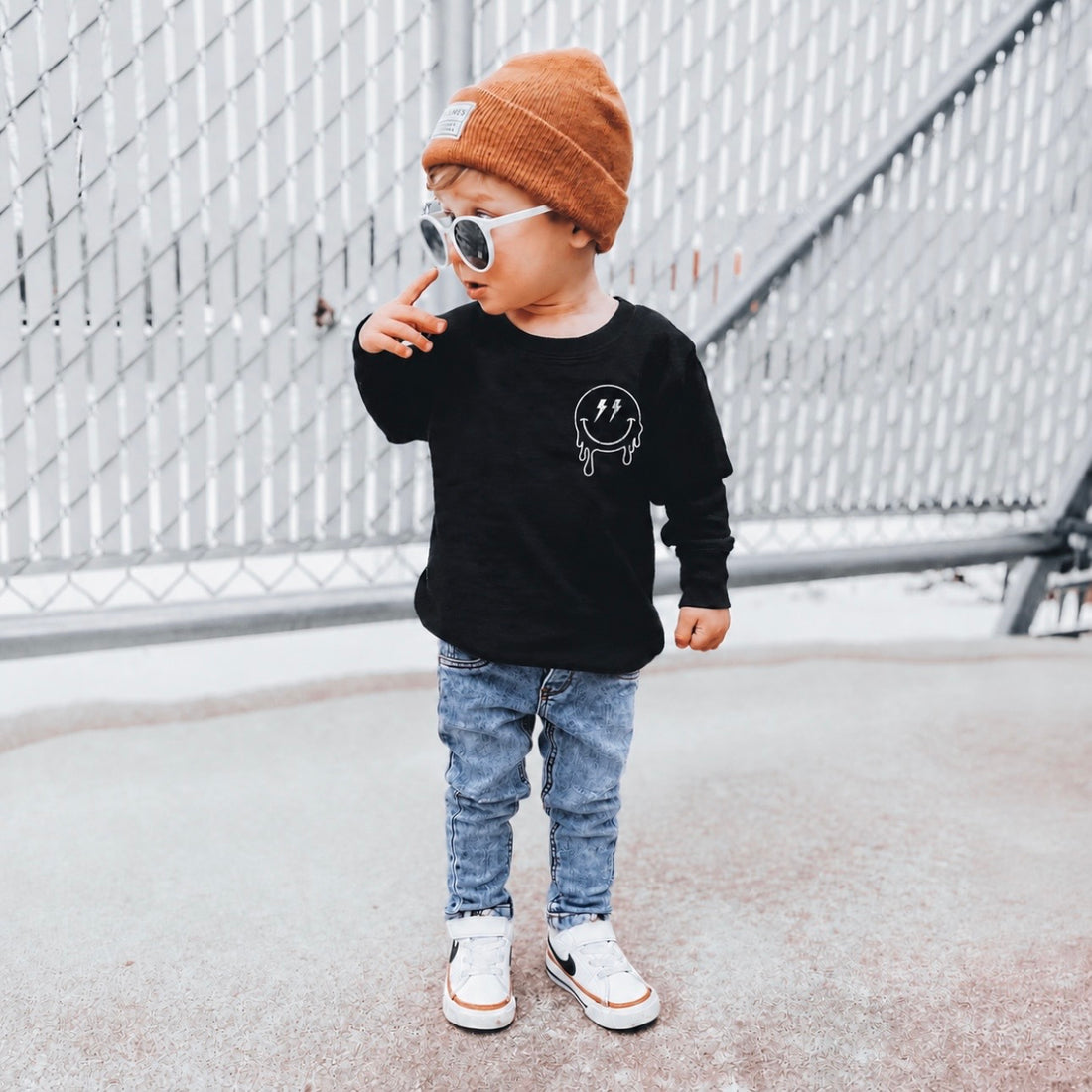 Toddler boy wearing LENOX JAMES classic midwash blue denim jeans with a slim fit, styled with a black smiley face graphic shirt, rust-colored beanie, white sunglasses, and sneakers while standing outdoors.