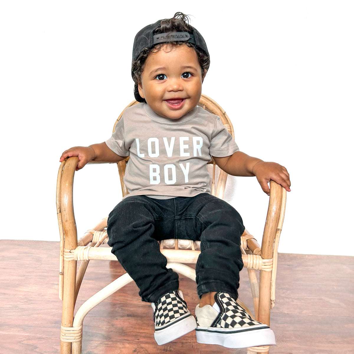 Toddler boy wearing LENOX JAMES black denim jeans styled with a taupe Lover Boy graphic T-shirt, backward black baseball cap, and checkerboard sneakers while sitting in a rattan chair.