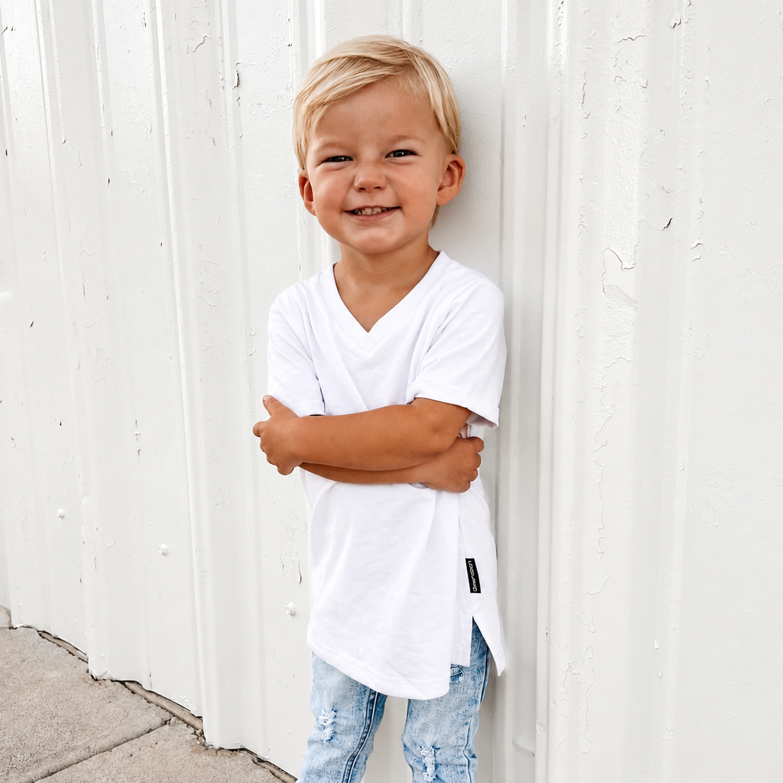A young blonde boy smiling and leaning against a white corrugated metal wall, wearing a white long-sleeve V-neck t-shirt and light blue jeans.