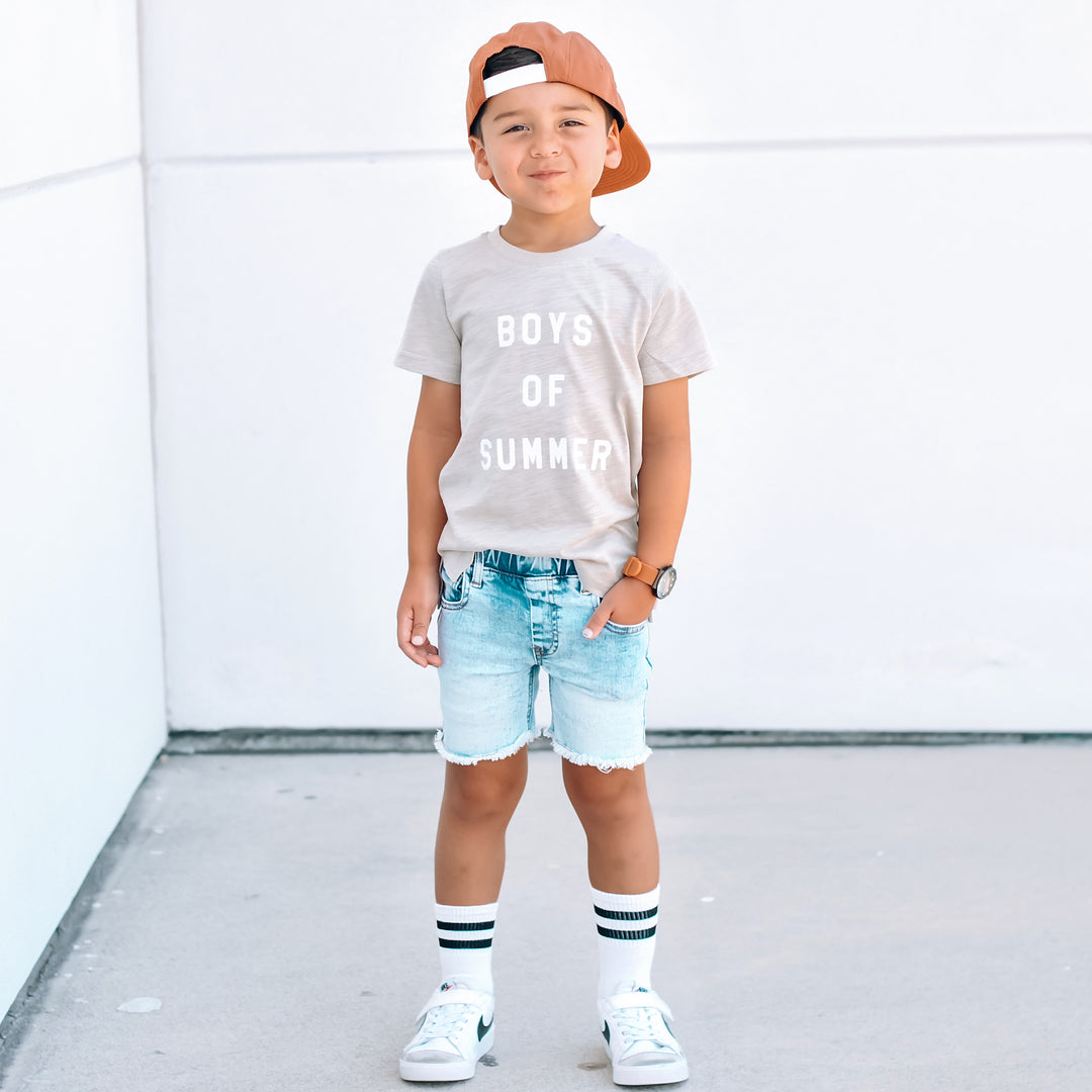 Child wearing a 'Boys of Summer' t-shirt, denim shorts, and a cap against a white background