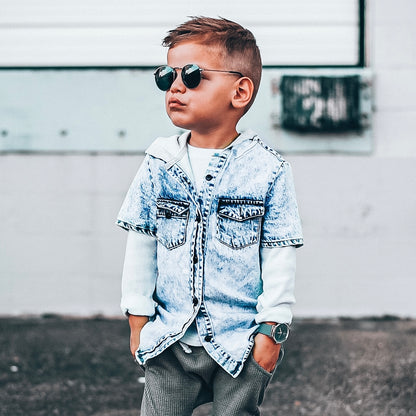 Child wearing sunglasses and a denim jacket in front of a white wall.