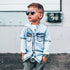 Child wearing sunglasses and a denim jacket in front of a white wall.