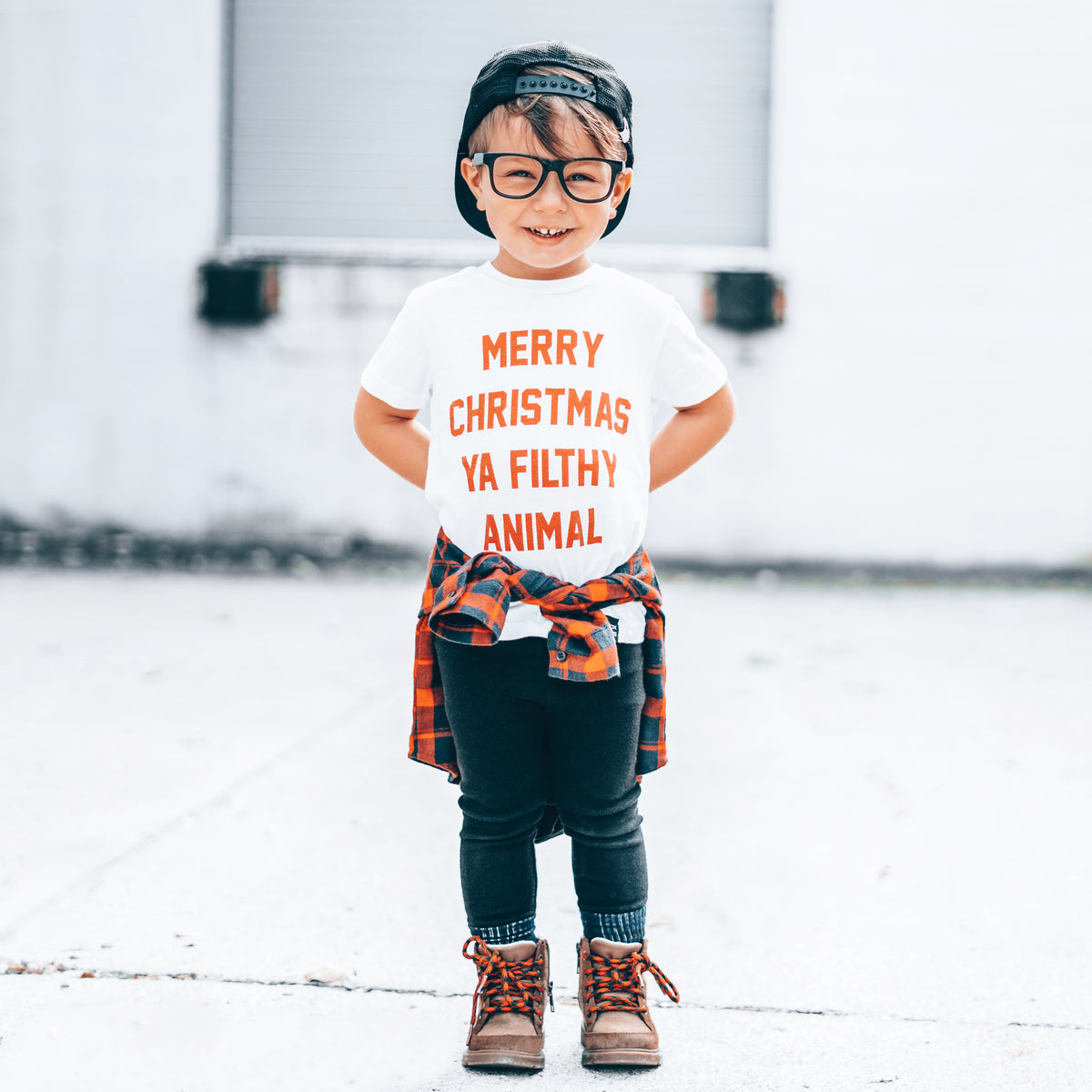 Child wearing LENOX JAMES Filthy Animal white Christmas graphic tee with red holiday lettering, black pants and plaid shirt tied at waist